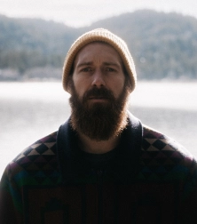 a man with a beard in a fishing outfit standing across a lake